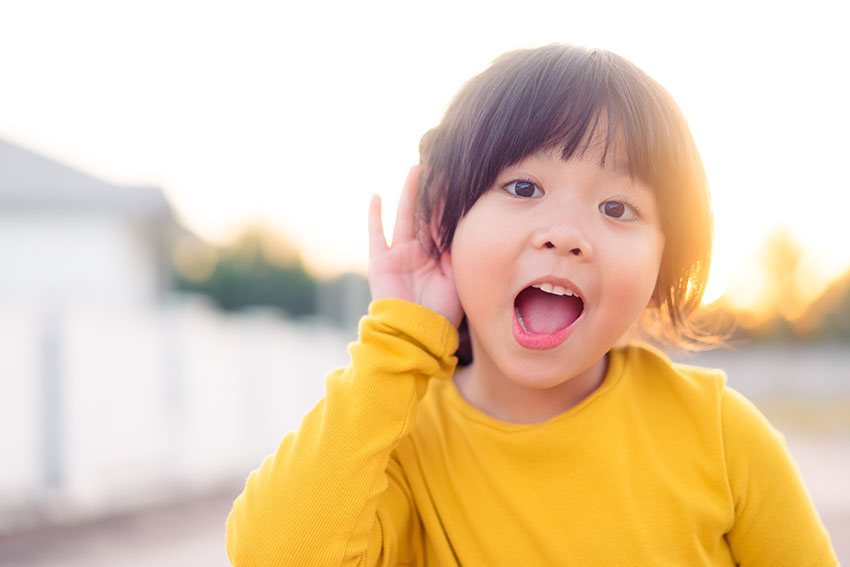 Liten asiatisk jente holder hånden nær øret og lytter. Spennende ansikt på asiatisk barn jente ha på seg gul skjorte om vinteren.